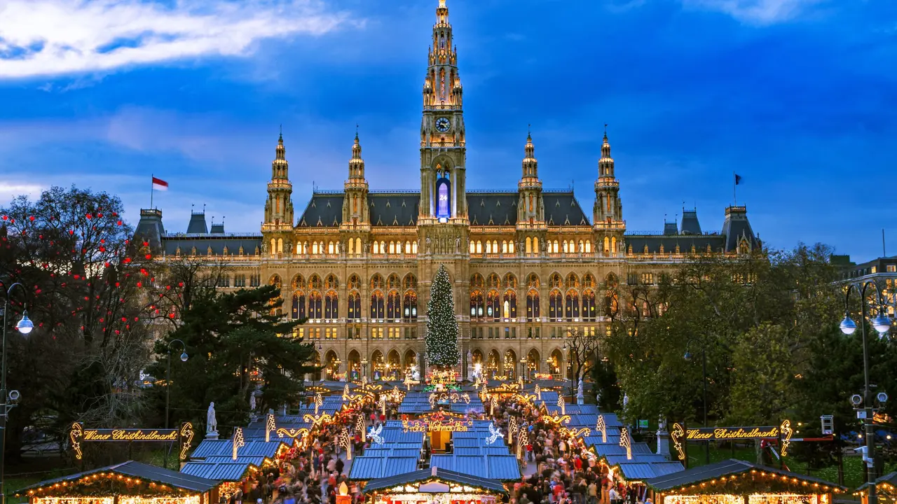 Shot of a gothic city hall which has a tall clock tower in the middle and four smaller towers, two either side, all with spiky turrets. The building has long windows all over and is a gold colour. Below are the Vienna Christmas markets, with four vertical strips of stalls, the centre having a christmas tree at the far end. There are many people shopping, with lights and lit up signs above them.