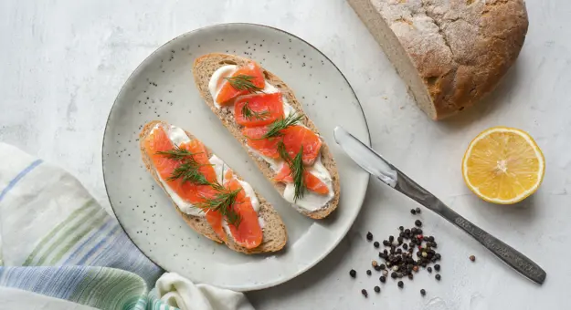 Gravalax on toast with cream cheese and dill, and a loaf of bread and lemon next to the plate