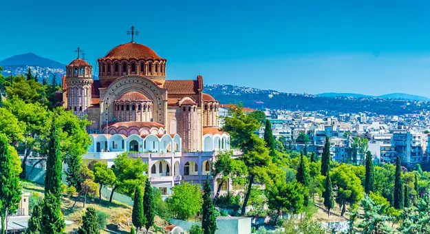 Brown and beige cathedral with a domed top with a cross on, trees in the forefront and a view of a town in the distance