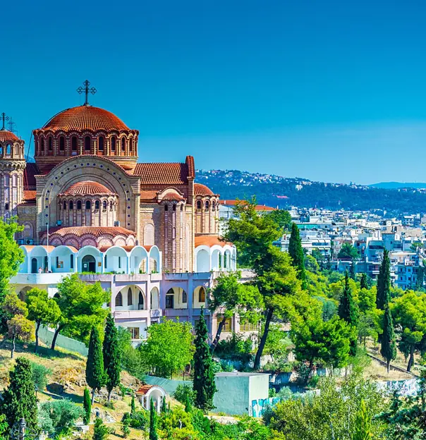 Brown and beige cathedral with a domed top with a cross on, trees in the forefront and a view of a town in the distance