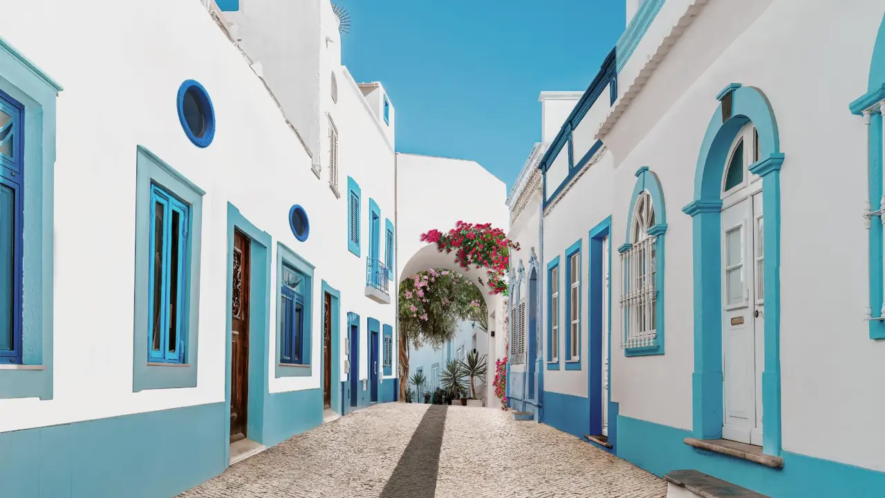 Blue and white buildings lining the cobbled streets of Olhão on the Algarve, with pink flowers adorning an archway at the end of the street