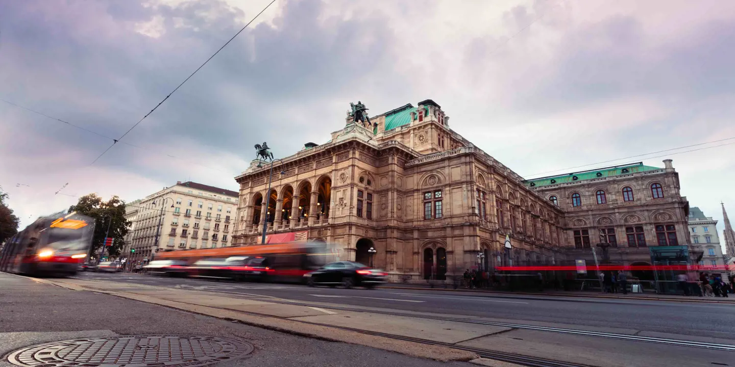 Low angle shot of Vienna Opera House, a beige coloured old-fashioned building with turquoise roofs and two statues of people on horses on either side of the front roof. Below is an action shot of vehicles moving, making them blurry, and a drain in the ground is in the left forefront. The sky is grey and cloudy.  