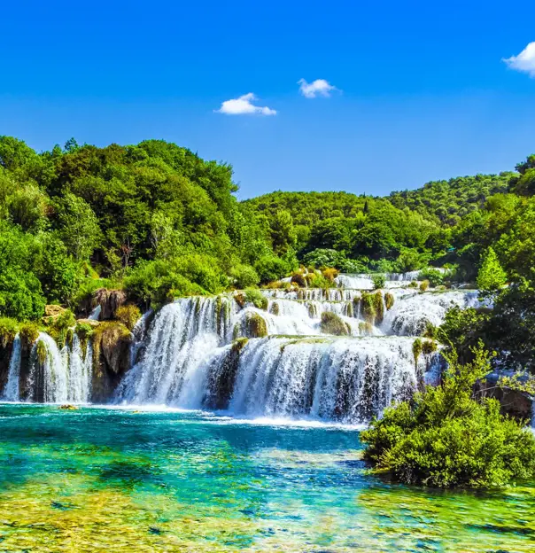 View of a waterfall in Krka National Park, with bright green trees behind it and a blue, clear sky above. Green and blue waters in the forefront.