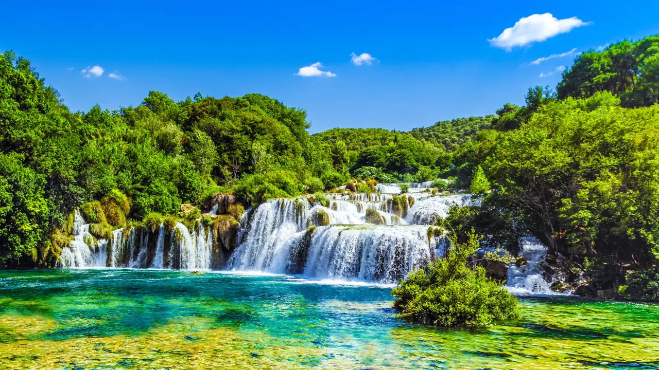 View of a waterfall in Krka National Park, with bright green trees behind it and a blue, clear sky above. Green and blue waters in the forefront.