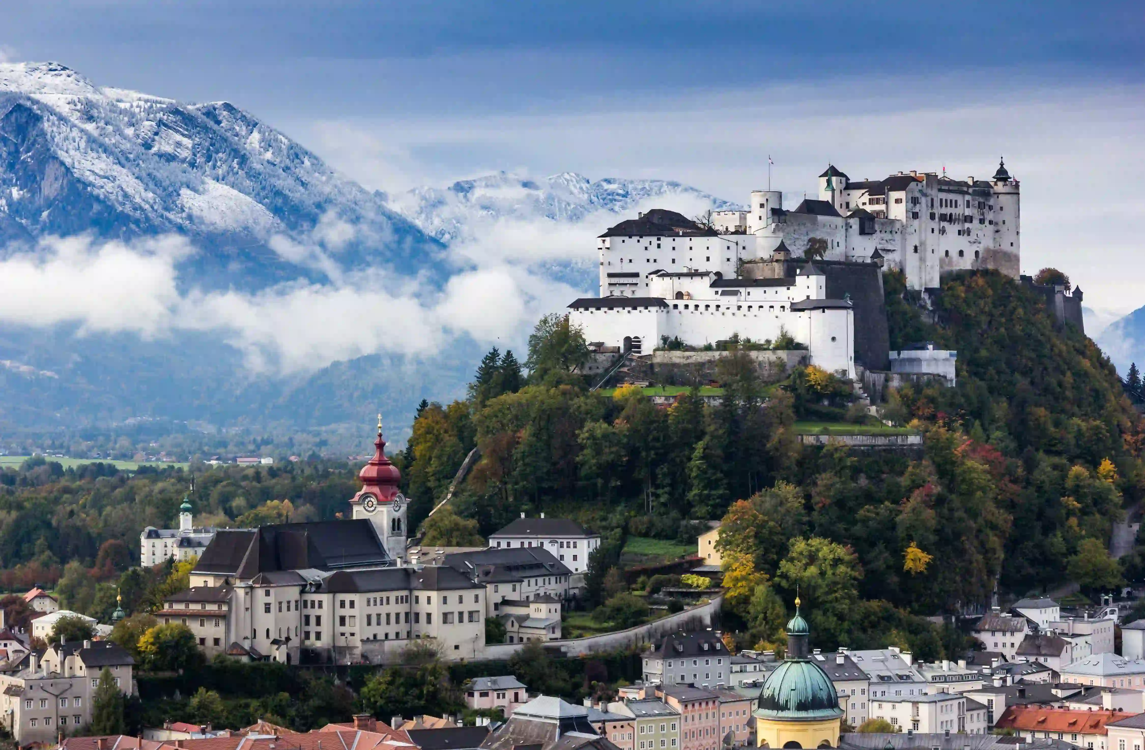 View of the Hohensalzburg Fortress, a white castle with black roofs on top of a grassy hill. At the bottom of the hill is a white large building with a red turret. Behind these is a large mountain with snow on and clouds in front of it. In the bottom right of the image is a snippet of the town, showing the roofs and the fronts of buildings.