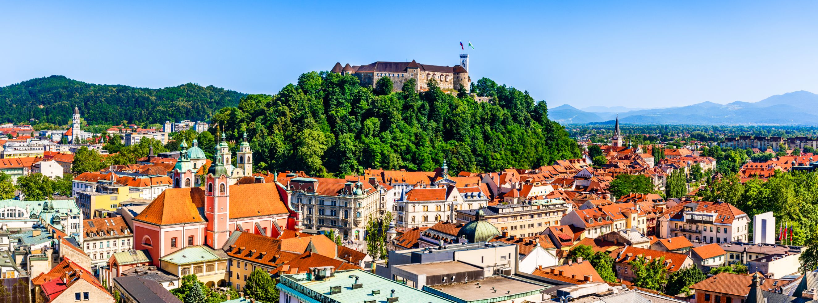 High angle shot of a town, showing lots of buildings with orange roofs and a hill in the centre covered in trees, with a large stone building on top. Mountains in the distance on the right and a mountain covered in trees on the left. 
