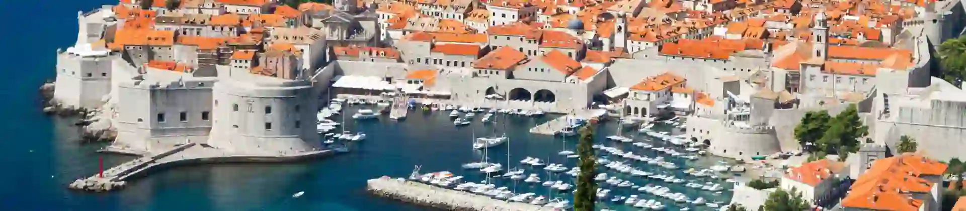 High angle view of Dubrovnik's coast and its harbour, with boats docked and lots of buildings with orange roofs behind. The navy blue sea to the left.