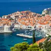 High angle view of Dubrovnik's coast and its harbour, with boats docked and lots of buildings with orange roofs behind. The navy blue sea to the left.