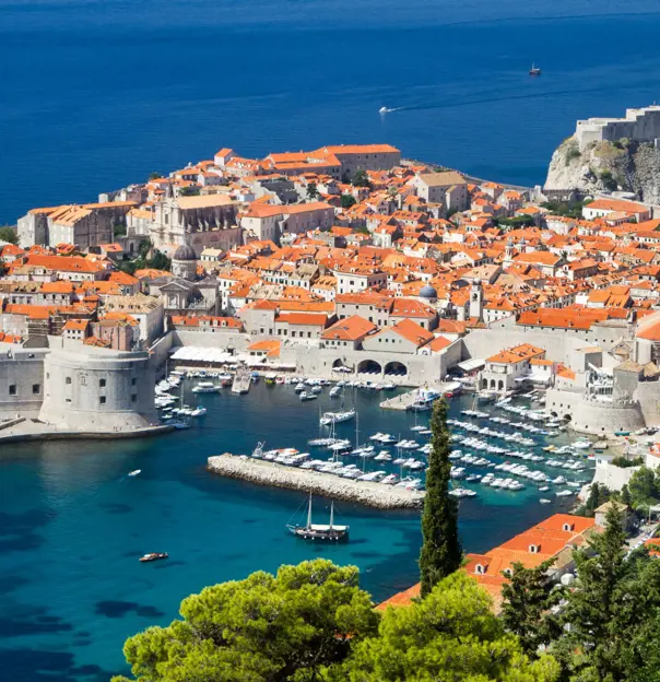 High angle view of Dubrovnik's coast and its harbour, with boats docked and lots of buildings with orange roofs behind. The navy blue sea to the left.