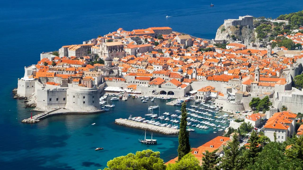 High angle view of Dubrovnik's coast and its harbour, with boats docked and lots of buildings with orange roofs behind. The navy blue sea to the left.