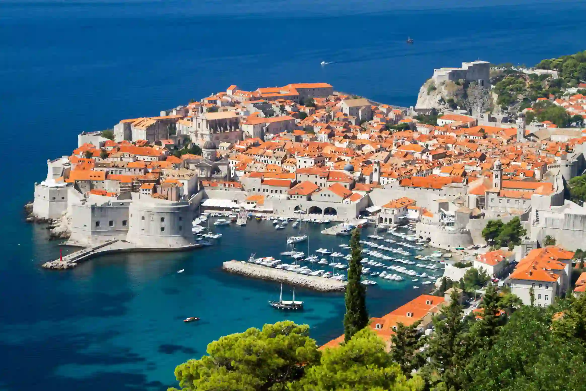 High angle view of Dubrovnik's coast and its harbour, with boats docked and lots of buildings with orange roofs behind. The navy blue sea to the left.