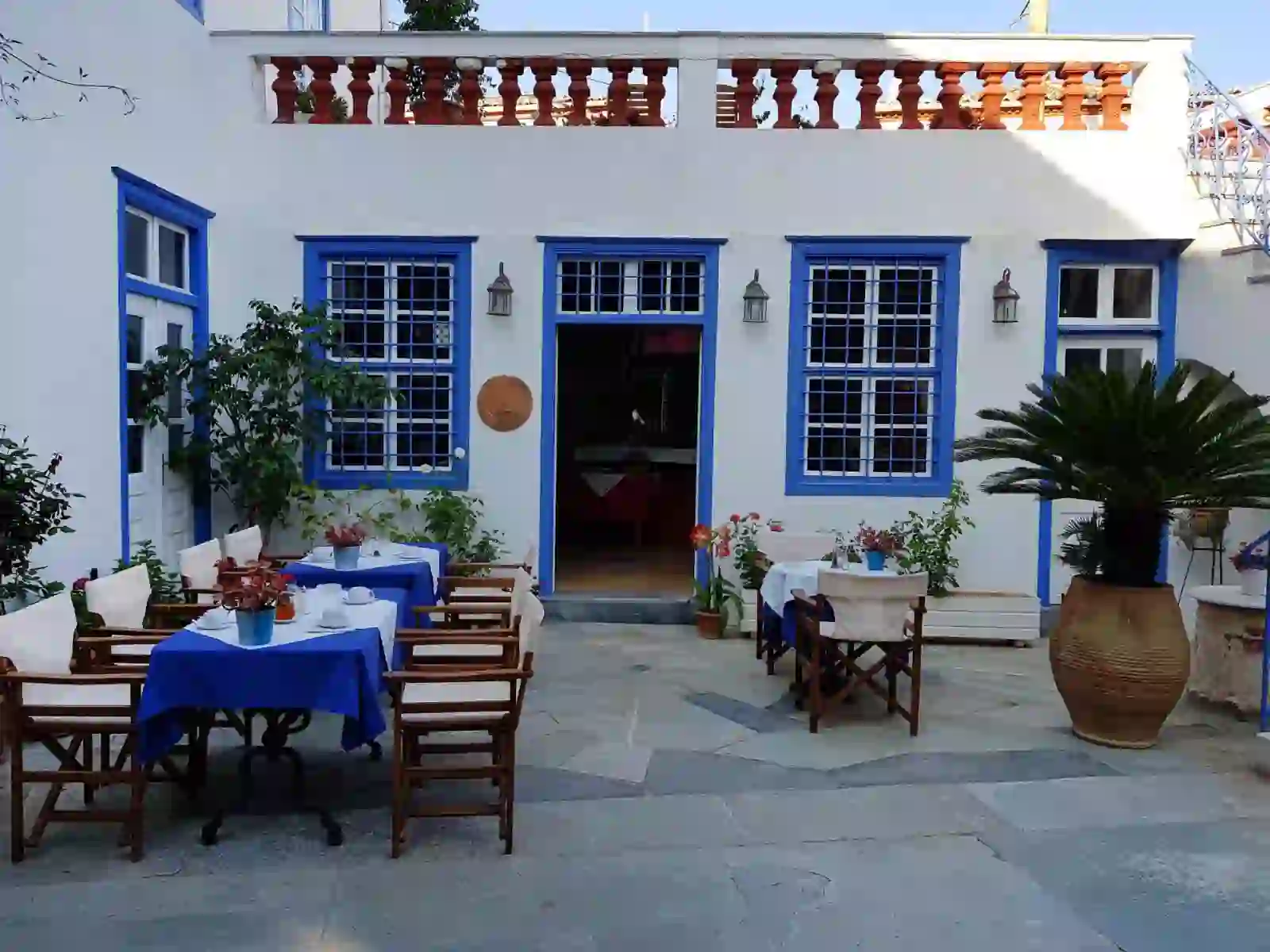 Outdoor dining area on a patio at Hotel Ippokampos in Hydra, Greece, featuring tables with blue tablecloths, surrounded by potted plants, with doors and windows framed in traditional blue paint
