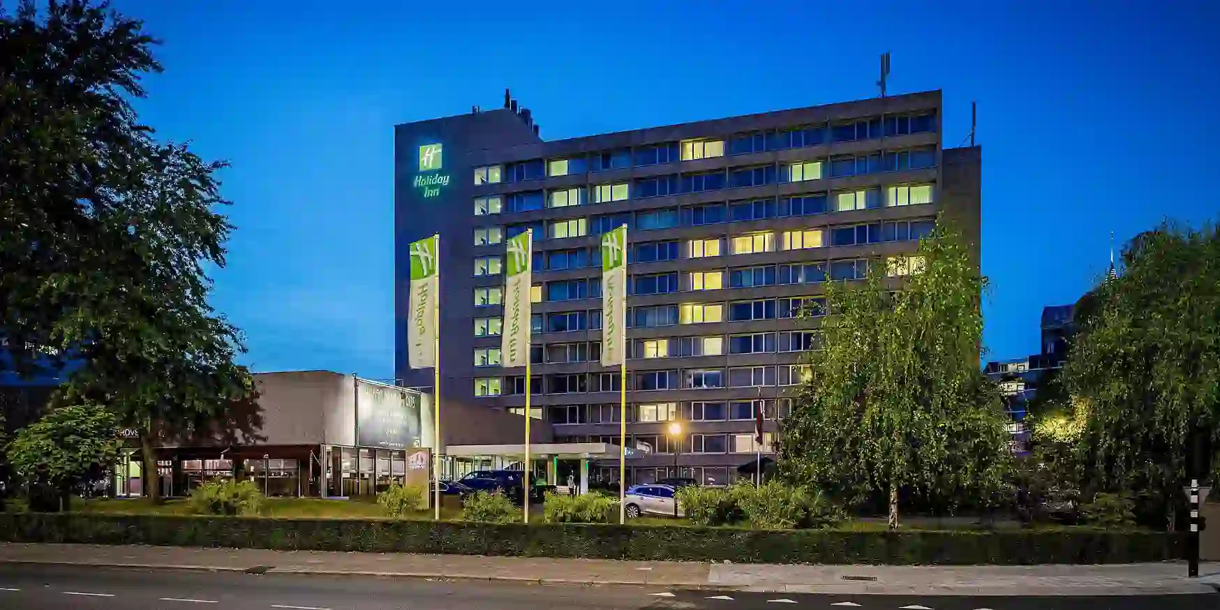 Exterior of the Holiday Inn Eindhoven, a modern building with signage on the top left, set beside a road with parked cars and green trees