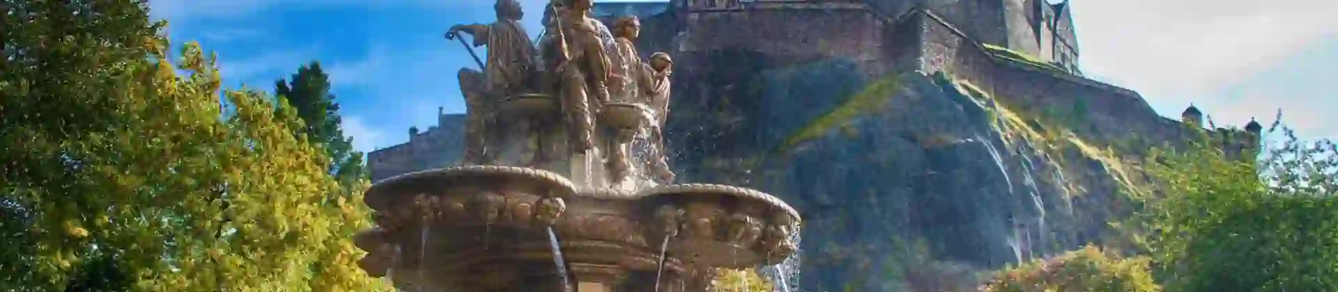 A view of Edinburgh Castle perched high on a rocky hill in the background, with a decorative fountain in the foreground below, surrounded by greenery