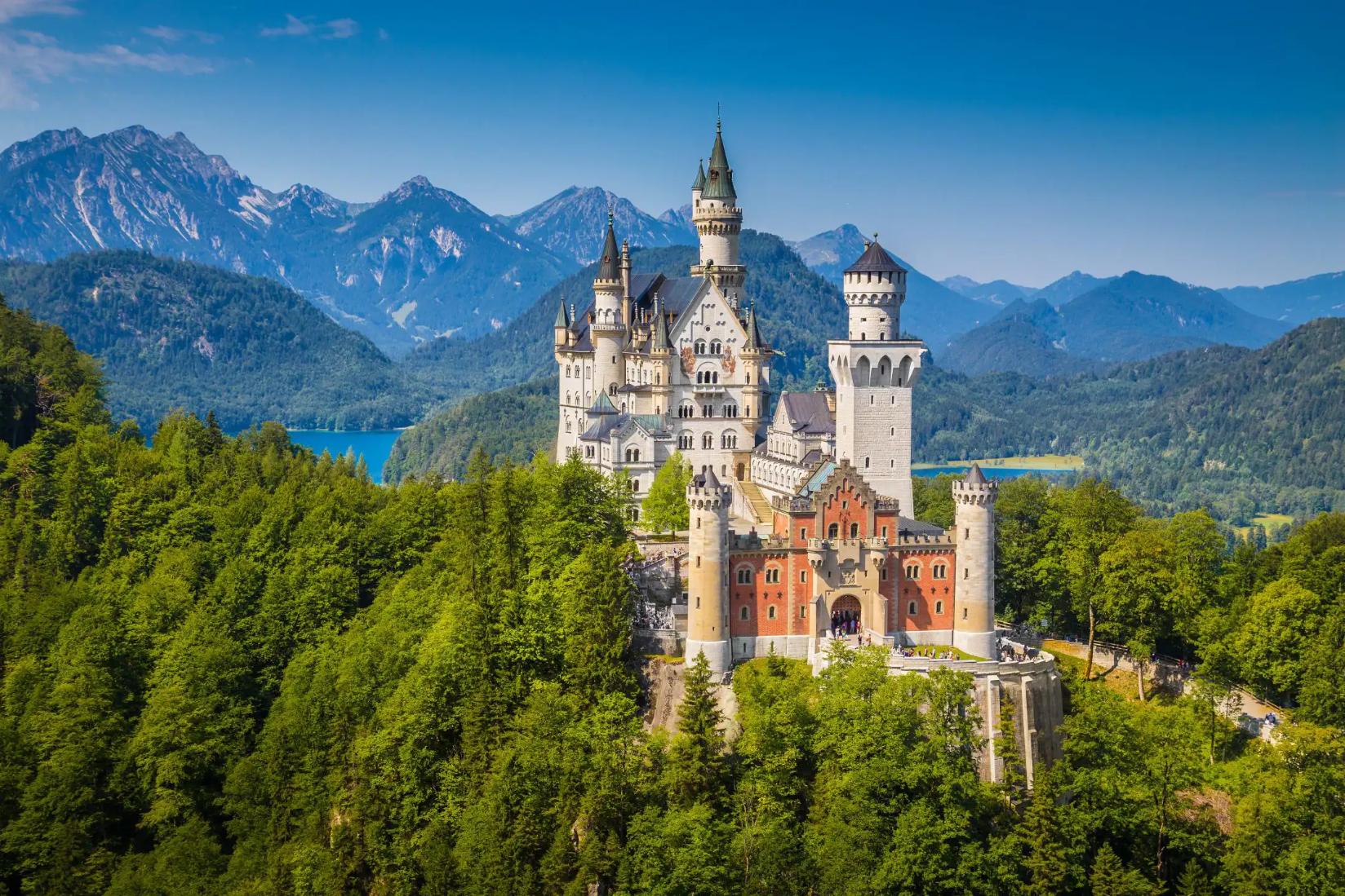 Neuschwanstein Castle in Bavaria, Germany, sitting atop a forested hill with mountain and a lake in the background