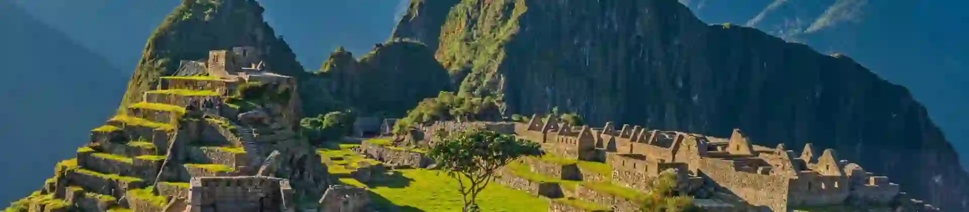 A panoramic view of Machu Picchu in Peru, showing ancient Incan ruins built into a green mountain ridge under a clear blue sky, with Huayna Picchu towering in the background