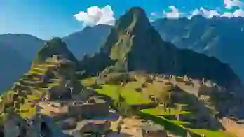 A panoramic view of Machu Picchu in Peru, showing ancient Incan ruins built into a green mountain ridge under a clear blue sky, with Huayna Picchu towering in the background