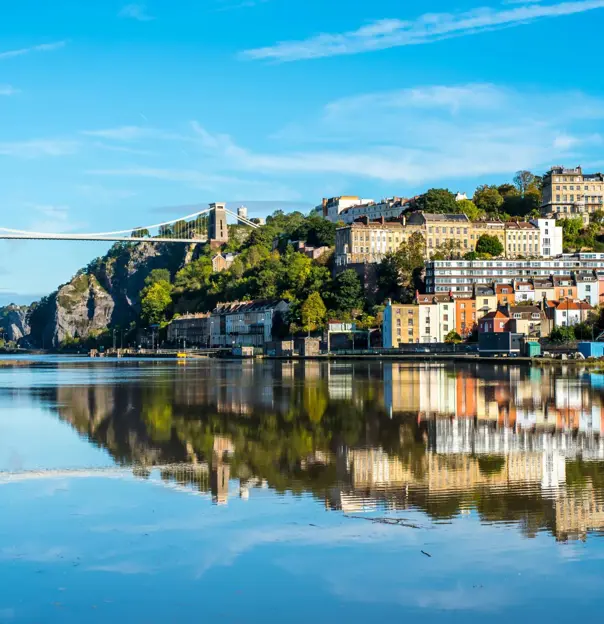 Clifton Suspension Bridge spanning the Avon Gorge in Bristol, with the River Avon in the foreground reflecting the bridge and the surrounding Clifton area under a clear sky