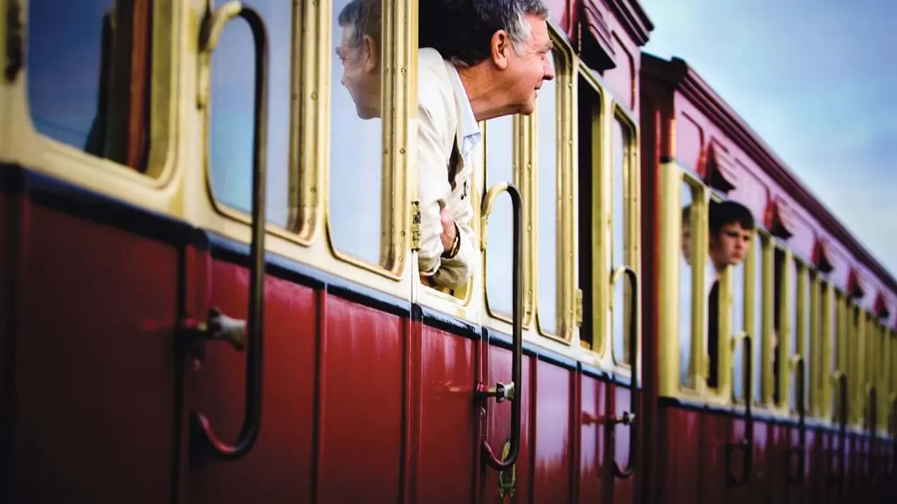 A man and boy poking their heads out of a window of a train in the Isle of Man, the man being further forward and in focus, and boy further away and blurry 