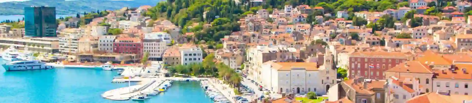 A panoramic view of Split, Croatia, showing the turquoise Adriatic Sea, a bustling harbour with yachts, and the terracotta rooftops of Old Town at the foot of lush green Marjan Hill