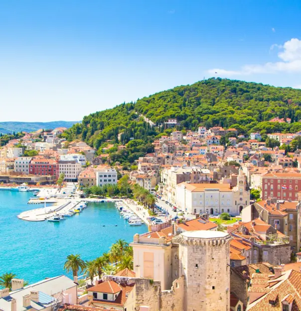 A panoramic view of Split, Croatia, showing the turquoise Adriatic Sea, a bustling harbour with yachts, and the terracotta rooftops of Old Town at the foot of lush green Marjan Hill