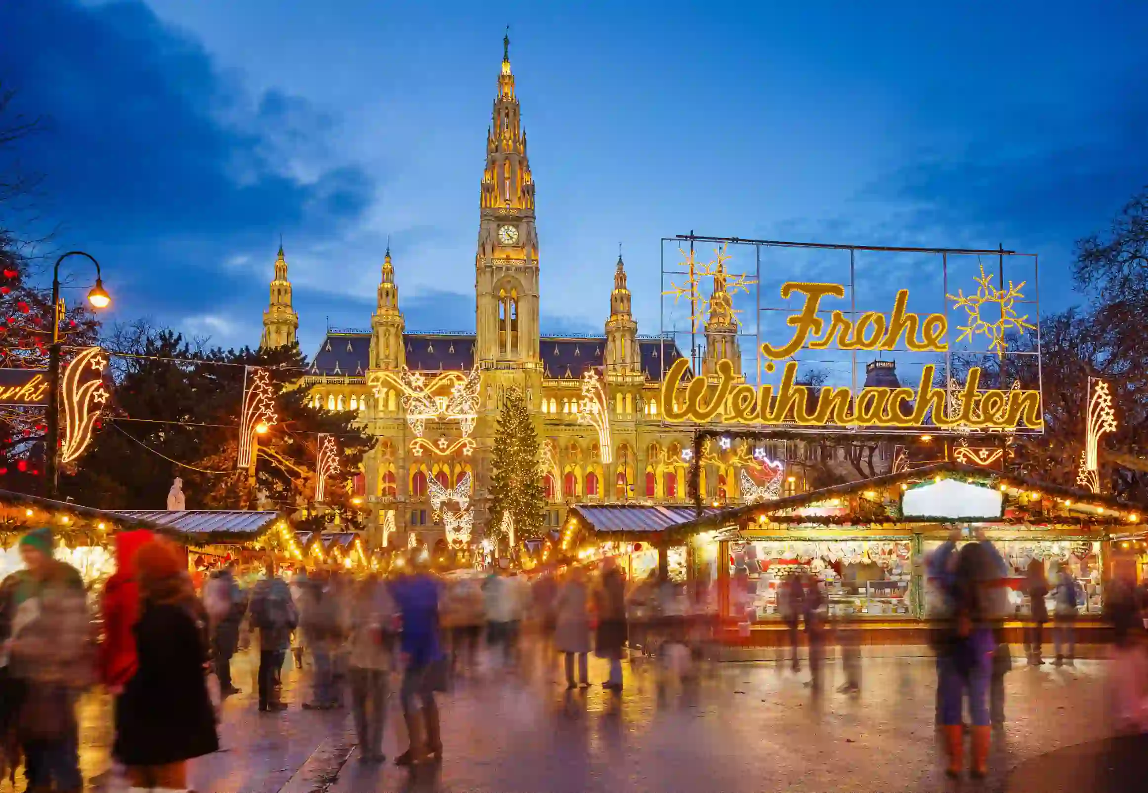 Shot of Vienna's gothic city hall which has one tall clock tower and four smaller towers, two on each side, all with pointy turrets. The building is a gold colour and is in front of a bright blue night sky. In front, is a Christmas Market, showing strips of cabin-like stalls and a variety of lit up Christmas decorations, one in the forefront reading 'Merry Christmas' in Austrian. People can be seen, although blurry as they are moving around. 