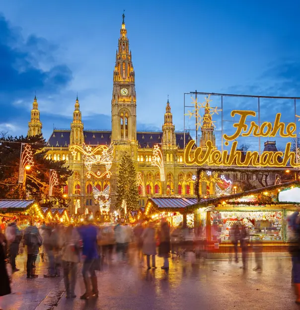 Shot of Vienna's gothic city hall which has one tall clock tower and four smaller towers, two on each side, all with pointy turrets. The building is a gold colour and is in front of a bright blue night sky. In front, is a Christmas Market, showing strips of cabin-like stalls and a variety of lit up Christmas decorations, one in the forefront reading 'Merry Christmas' in Austrian. People can be seen, although blurry as they are moving around.