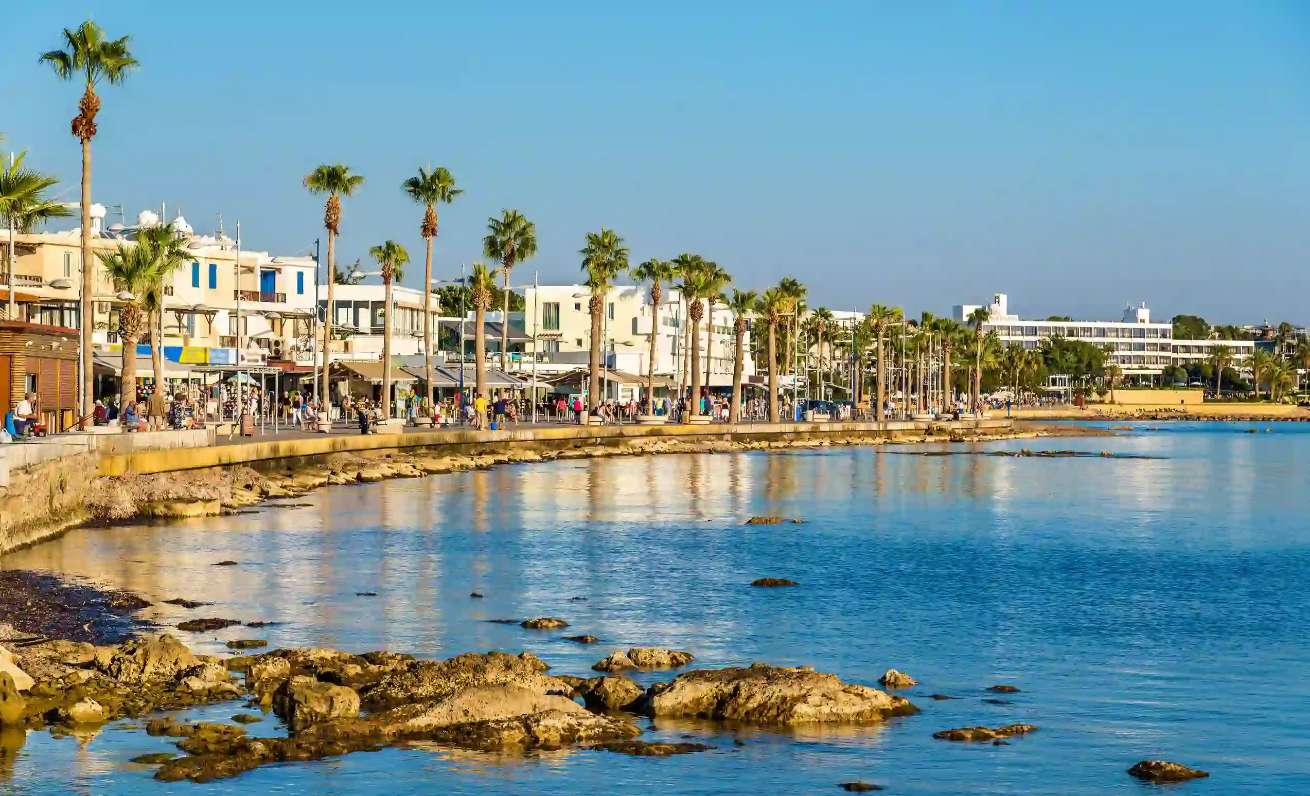 View of a coastal town, with the sea and a line of palm trees lining the edge of the land, behind them, people and shops and restaurants. The sky is clear and blue.