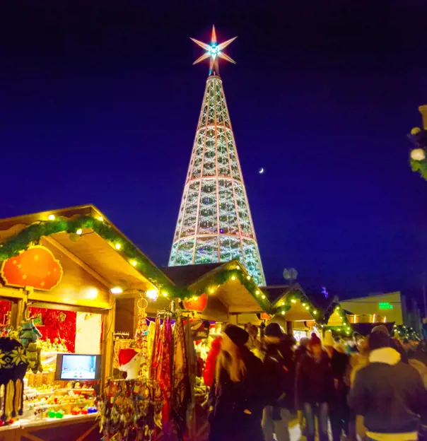 View of a Christmas market, with wooden, cabin-like stalls in the forefront, selling winter hats, dream catchers and scarves. The roofs are lined with green leaf-like garlands and fairy lights. Behind these is a Christmas tree metal structure, with a large, lit up star on top, in front of a navy blue night sky.