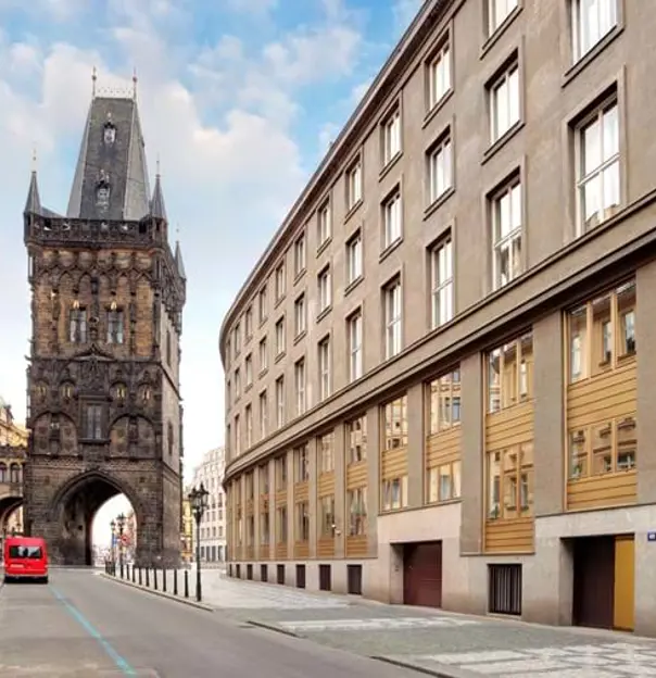 Shot of a street in Prague, buildings either side, and red car at the end, heading towards the archway of a dark-coloured tower with turrets. 