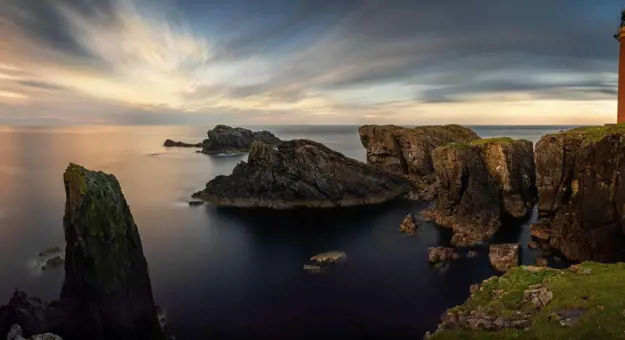 Long shot of the coast of the Isle of Lewis at sunset, showing the Butt of Lewis Lighthouse on the far right, looking out to sea