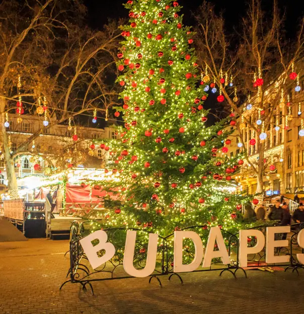 A square in Budapest at Christmas, in the front, a 'Budapest' sign, with a Christmas tree with lights and red baubles behind. Behind this, lit up decorations with hanging baubles, strung across the square.