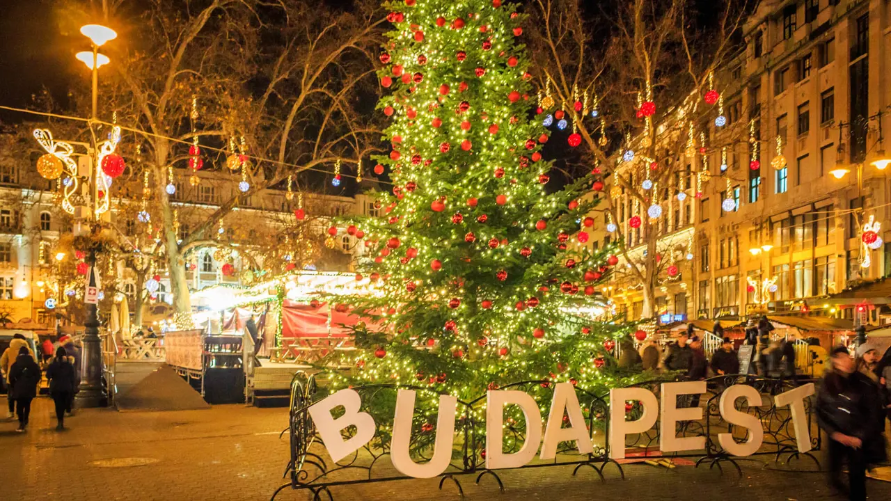 A square in Budapest at Christmas, in the front, a 'Budapest' sign, with a Christmas tree with lights and red baubles behind. Behind this, lit up decorations with hanging baubles, strung across the square.