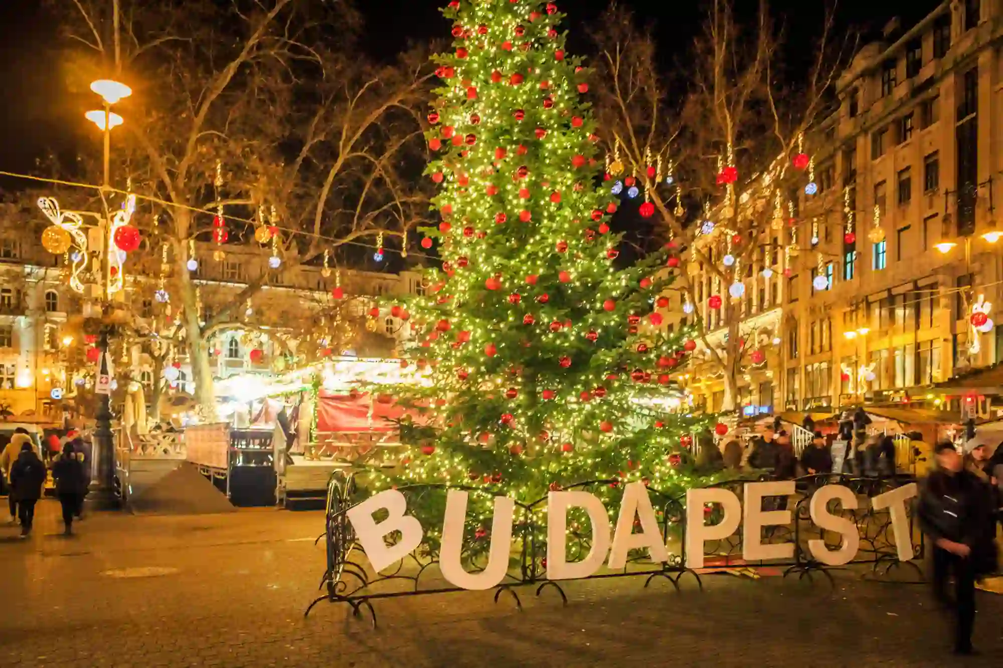 A square in Budapest at Christmas, in the front, a 'Budapest' sign, with a Christmas tree with lights and red baubles behind. Behind this, lit up decorations with hanging baubles, strung across the square.