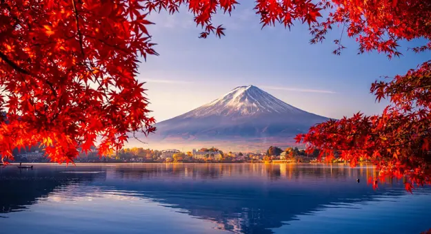Mount Fuji reflected in Lake Kawaguchi, framed by vivid red autumn leaves, with a sunlit lakeside town between the water and the mountain
