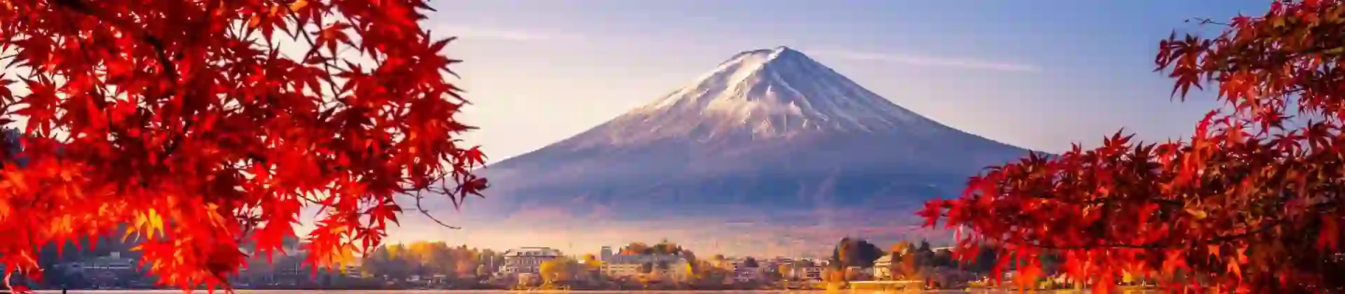 Mount Fuji reflected in Lake Kawaguchi, framed by vivid red autumn leaves, with a sunlit lakeside town between the water and the mountain