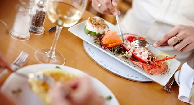 Image of the table with two people having dinner, from the point of view from behind one's shoulder, the plate in the forefront is blurry but the other shows a plate of lobster and vegetables, and the persons' two hands cutting it up with a knife and fork. A glass of white wine and salt and pepper grinders are also on the table.