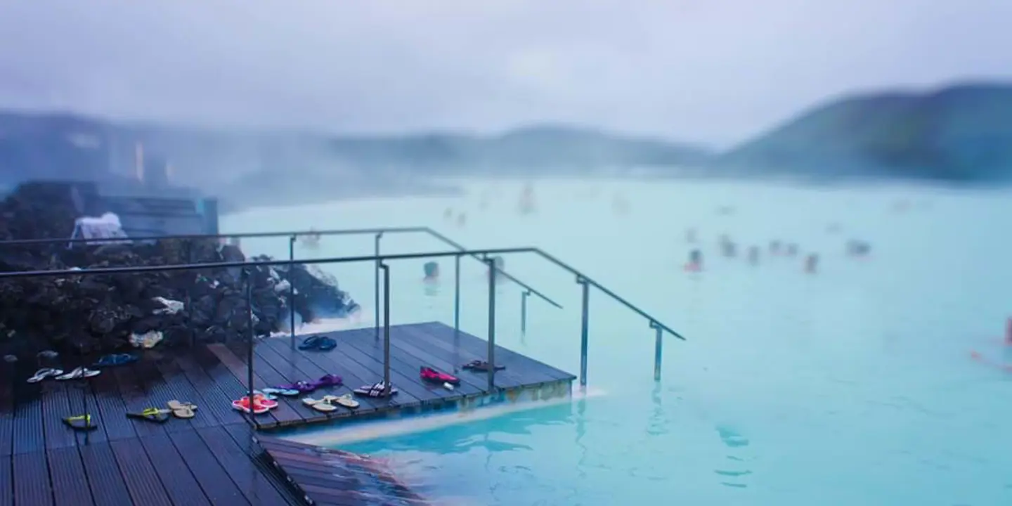 Geothermal lagoon in Iceland - bright blue water with steam coming off, mist clouding the view. In the forefront, a wooden platform descending into the water, with pairs of flip flops.