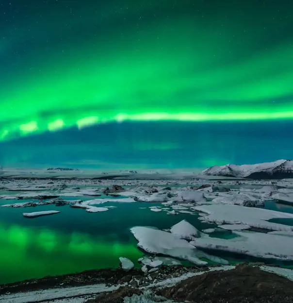 Green northern lights curving from the right back to the left. Below, a lake with ice blocks