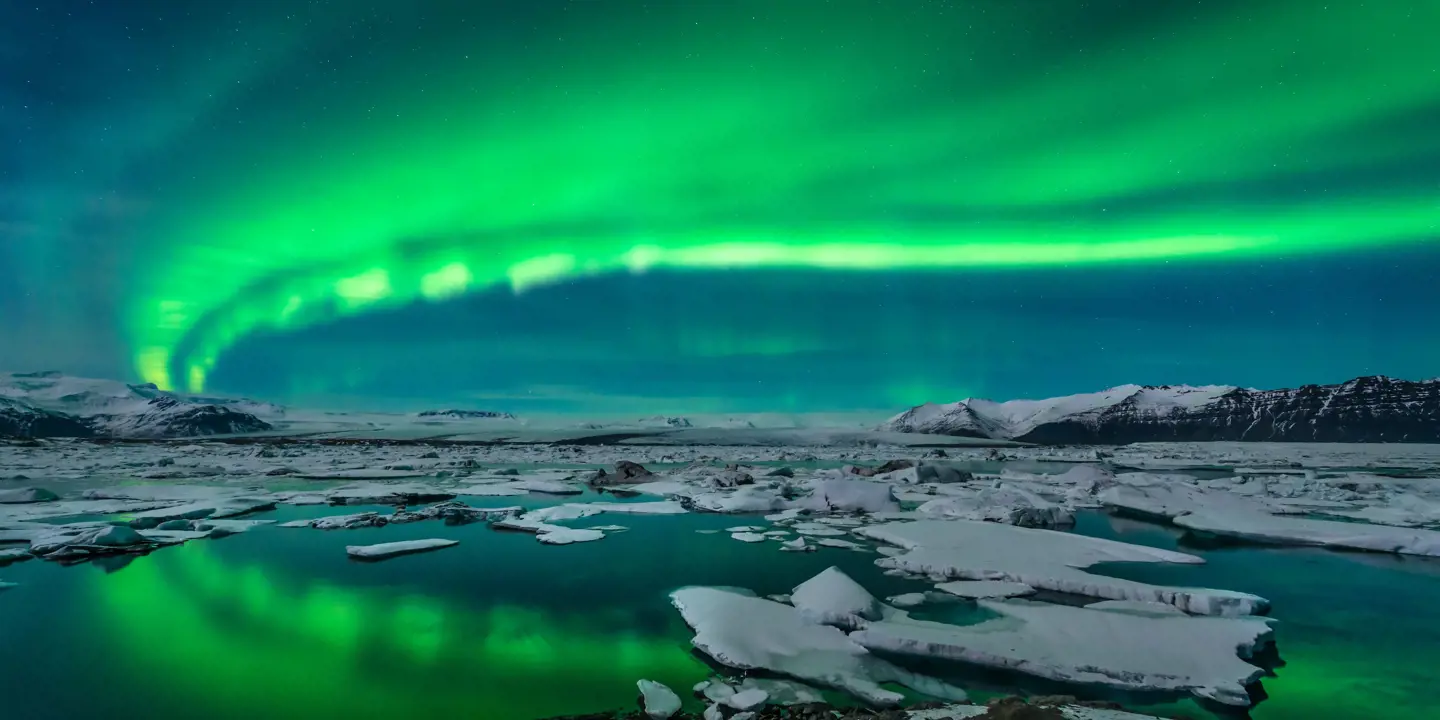 Green northern lights curving from the right back to the left. Below, a lake with ice blocks