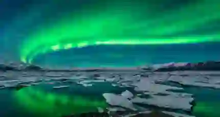 Green northern lights curving from the right back to the left. Below, a lake with ice blocks