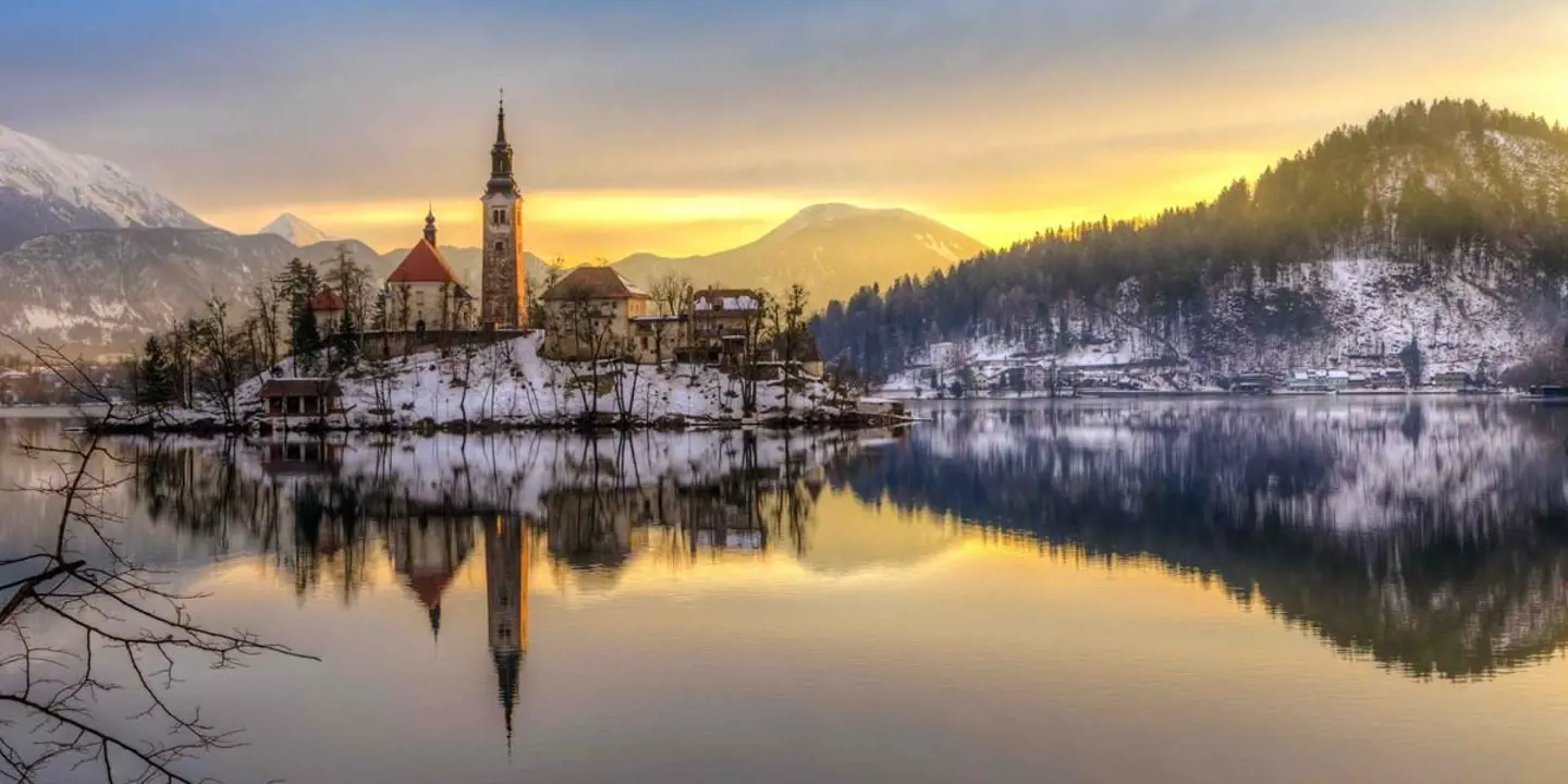 A lake at winter time, with a church in the centre with a slim tower and spiked turret. Mountains in the distance