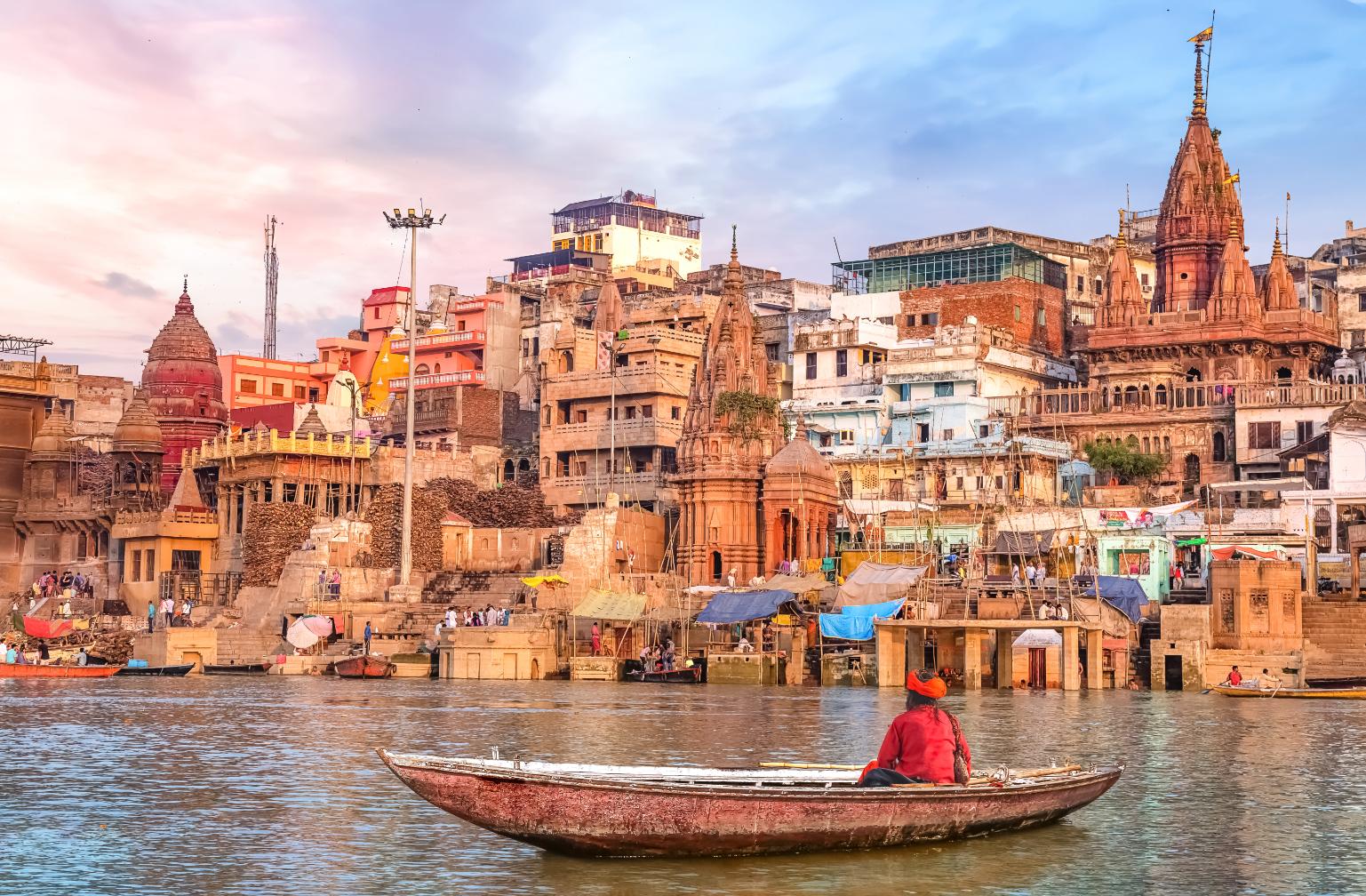 A boatman rowing on the River Ganges in Varanasi, facing the colourful temples and crowded ghats along the ancient city’s waterfront under a soft pastel sky