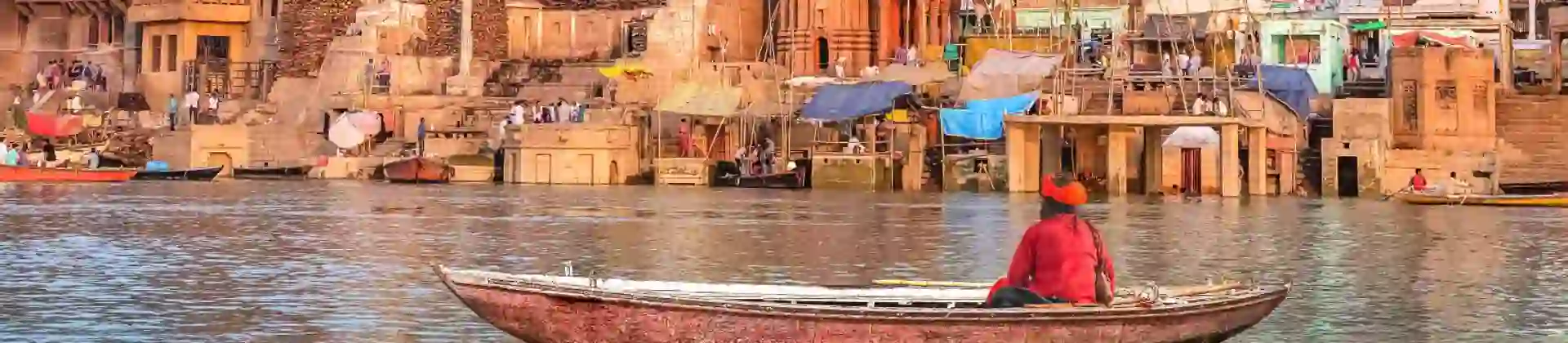 A boatman rowing on the River Ganges in Varanasi, facing the colourful temples and crowded ghats along the ancient city’s waterfront under a soft pastel sky