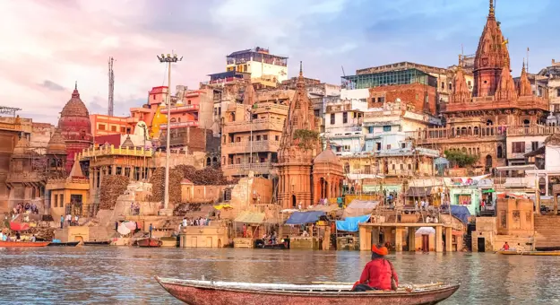 A boatman rowing on the River Ganges in Varanasi, facing the colourful temples and crowded ghats along the ancient city’s waterfront under a soft pastel sky