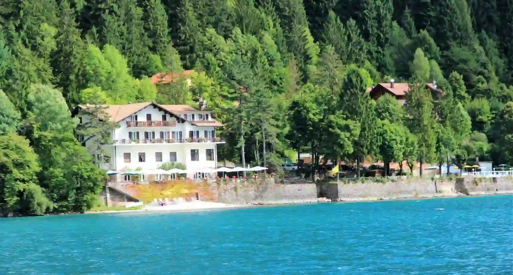 View of Lago Park Hotel on the shore of Lake Molveno, with the calm blue lake in the foreground and a forested mountain landscape in the background