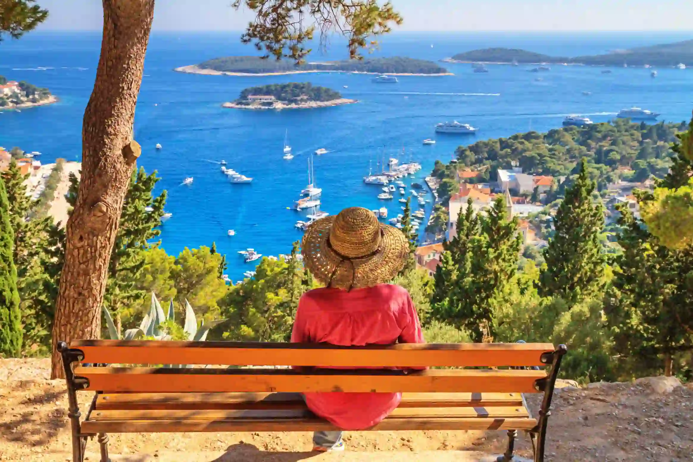 Back of a woman wearing a sun hat, sat on a bench looking out to sea. Variety of boats on the water, and forested land to the right.