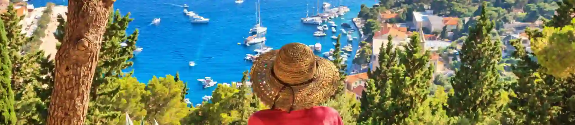 Back of a woman wearing a sun hat, sat on a bench looking out to sea. Variety of boats on the water, and forested land to the right.