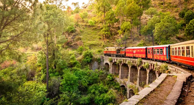 The heritage toy train crossing a railway bridge on the Kalka–Shimla route, surrounded by lush green hills and trees