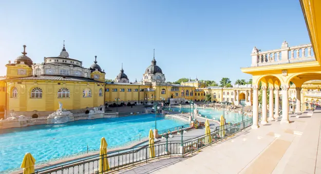 Szechenyi Thermal Baths, showing one large swimming pool and a smaller one to the right. Behind, a yellow building with grey domes and turrets on top.